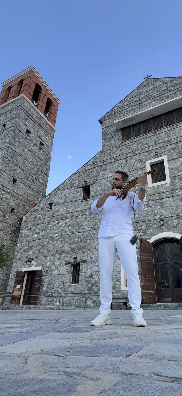 Marios playing violin in front of a stone church with a bell tower