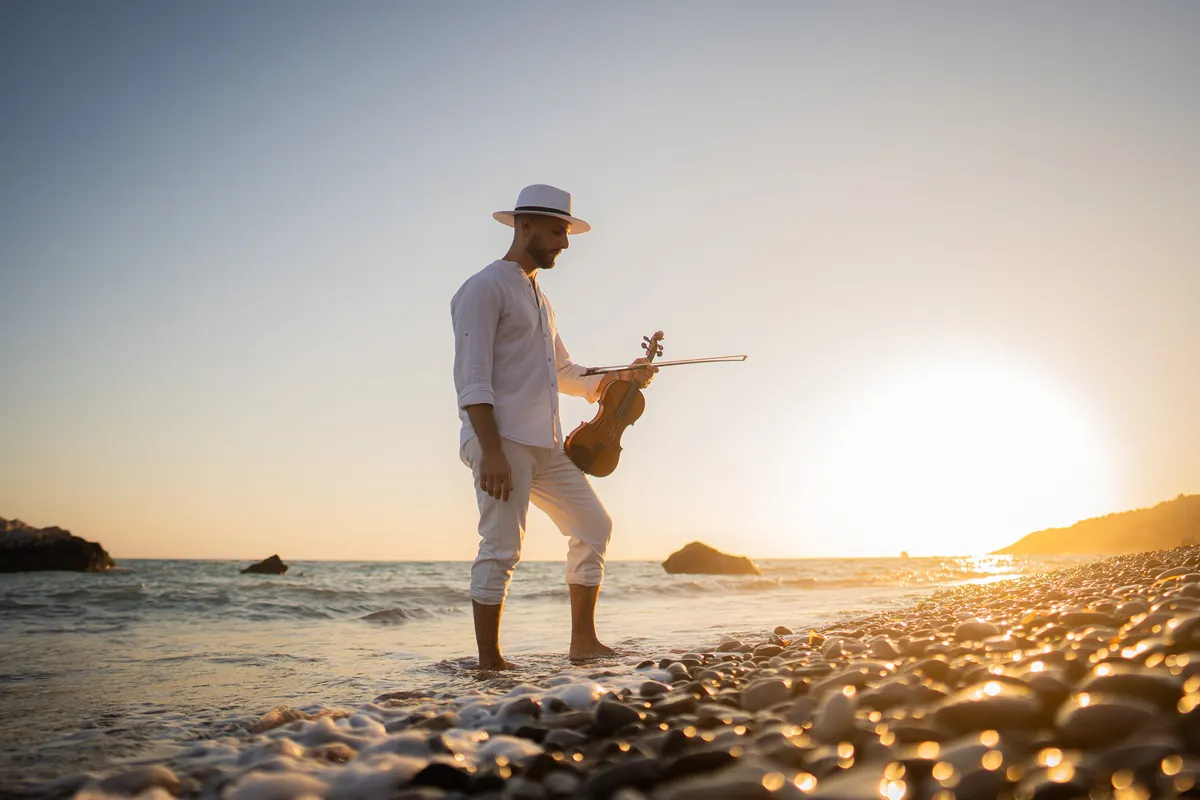 Marios in a white hat holding his violin on a beach at sunset