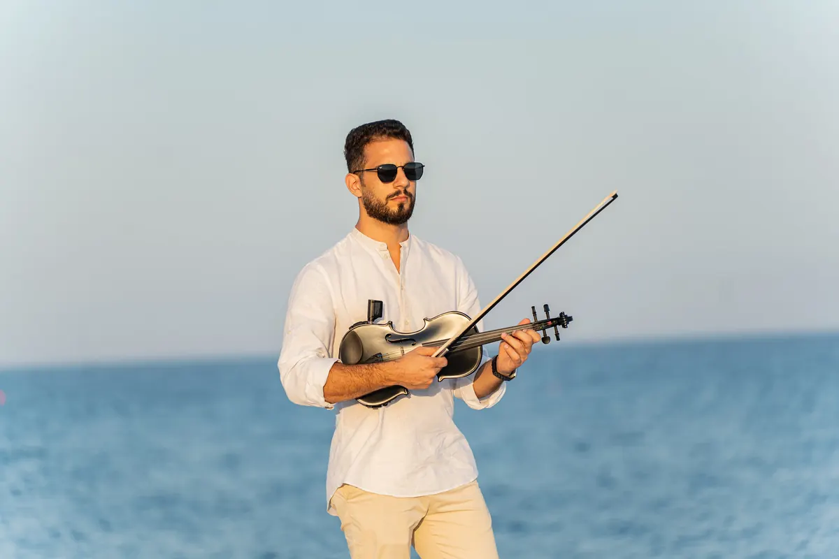 Marios in sunglasses holding a black electric violin by the sea