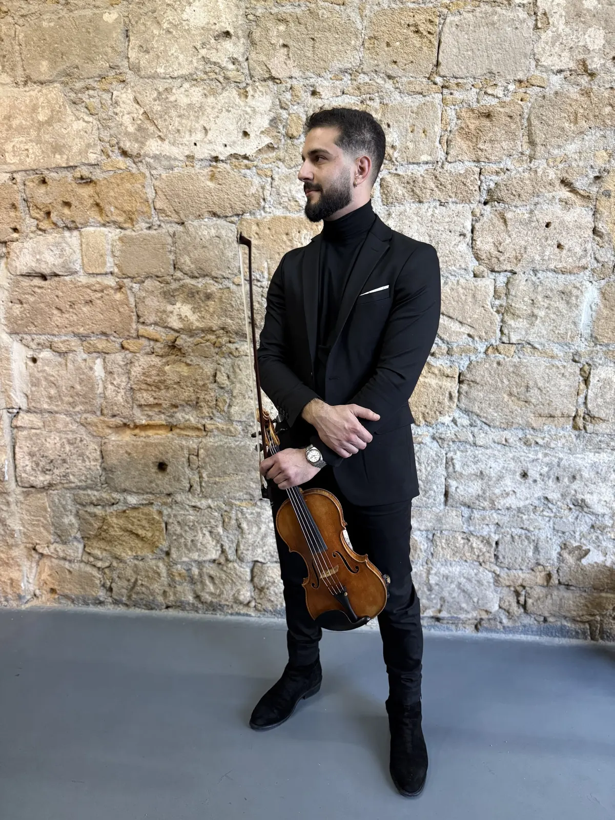 Marios in a black suit holding his violin in front of a stone wall
